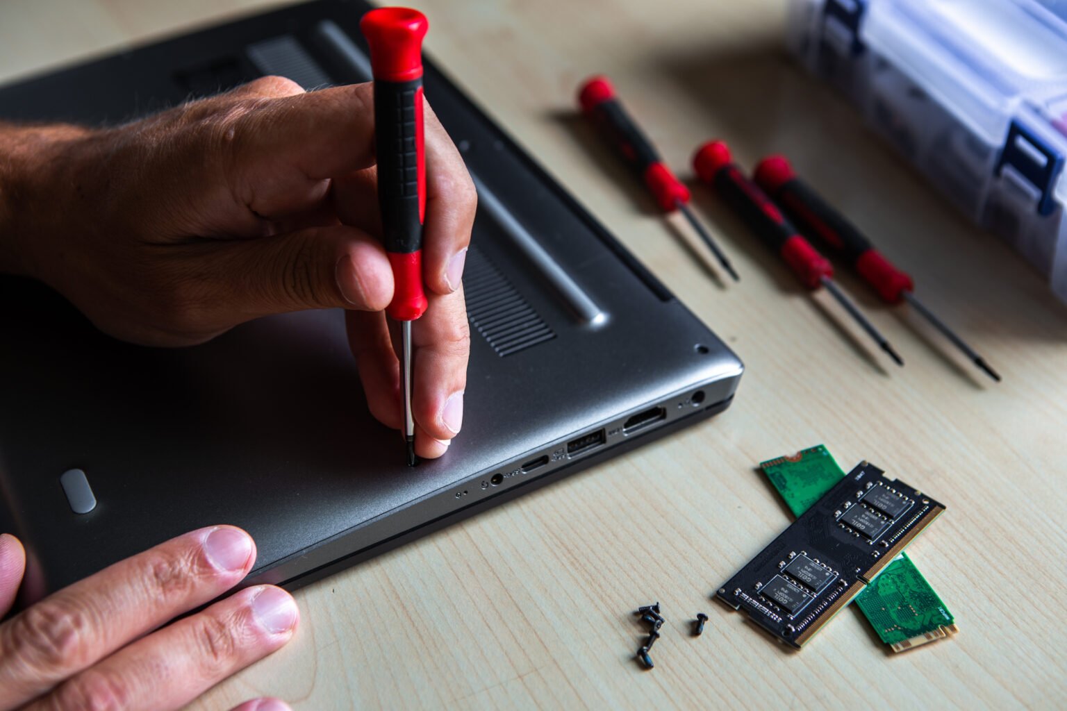 The hand of a young serviceman man opening a laptop computer with a red screwdriver. the laptop is on the desktop and next to it are the ssd disk and the ram memory.